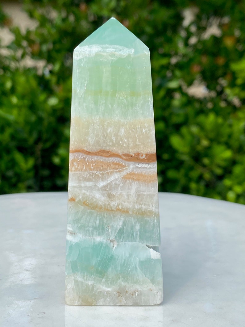 May include: A green, white, and brown banded onyx obelisk crystal point. The crystal is polished and has a smooth, glassy finish.