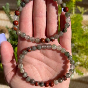 Pu&ograve; includere: Due braccialetti fatti con perline di pietre preziose verdi, grigie e rosse. I braccialetti sono impilati uno sopra l'altro.