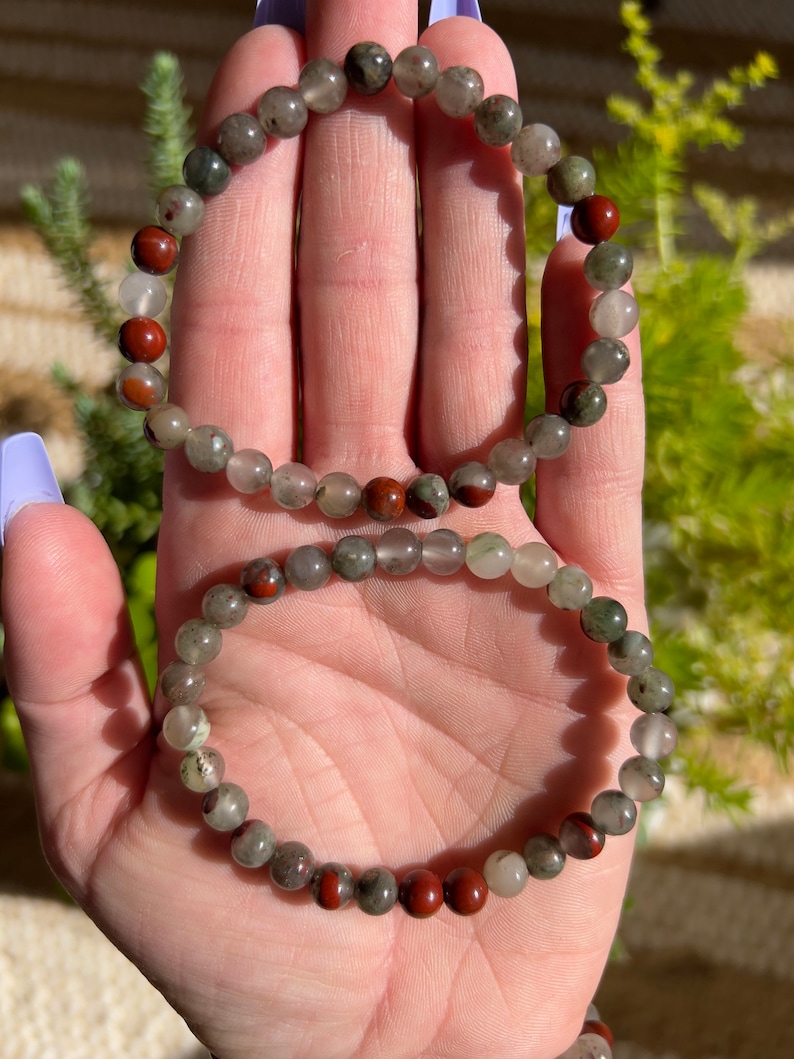 Pu&ograve; includere: Due braccialetti fatti con perline di pietre preziose verdi, grigie e rosse. I braccialetti sono impilati uno sopra l'altro.