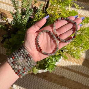 Pu&ograve; includere: Due braccialetti fatti con perline di pietre preziose verdi, grigie e rosse. I braccialetti sono impilati sul polso di una persona.