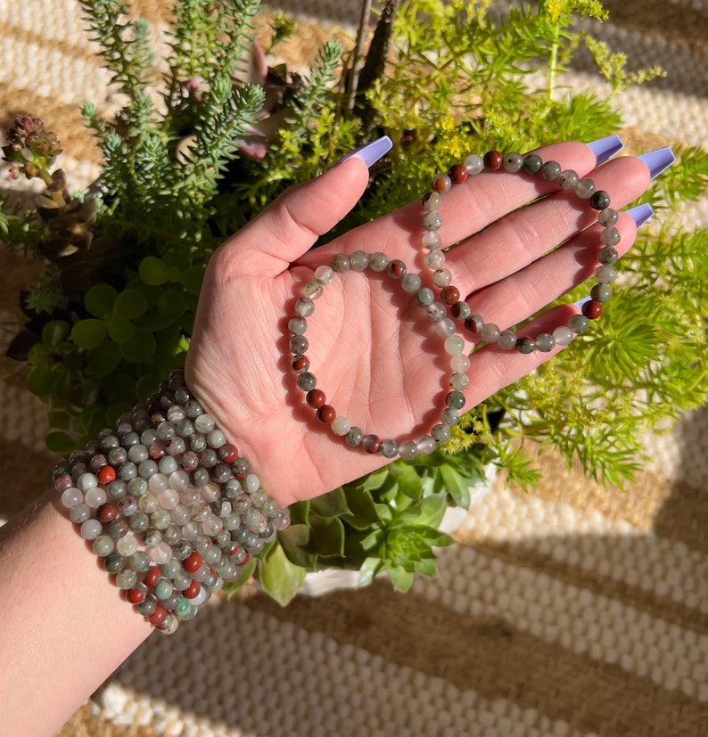 Pu&ograve; includere: Due braccialetti fatti con perline di pietre preziose verdi, grigie e rosse. I braccialetti sono impilati sul polso di una persona.