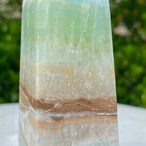 May include: A green, white, and brown layered obelisk crystal point. The crystal has a smooth, polished surface and is a natural stone.