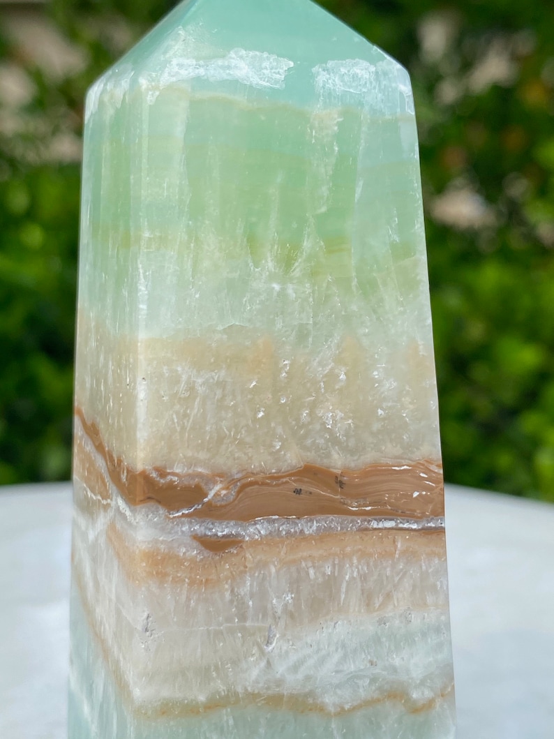 May include: A green, white, and brown layered obelisk crystal point. The crystal has a smooth, polished surface and is a natural stone.