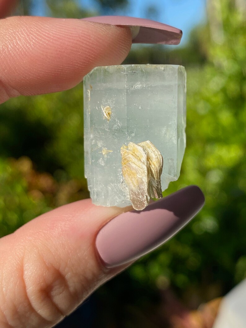 May include: A light blue aquamarine crystal with a clear, glassy texture. The crystal has a natural, rough shape and is accented with a small, golden-brown mineral inclusion.