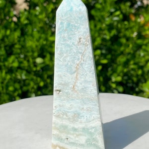 May include: A light blue and white celestite crystal point, also known as a tower or obelisk, with a pointed top and a flat base. The crystal has a natural, raw, and unpolished finish.