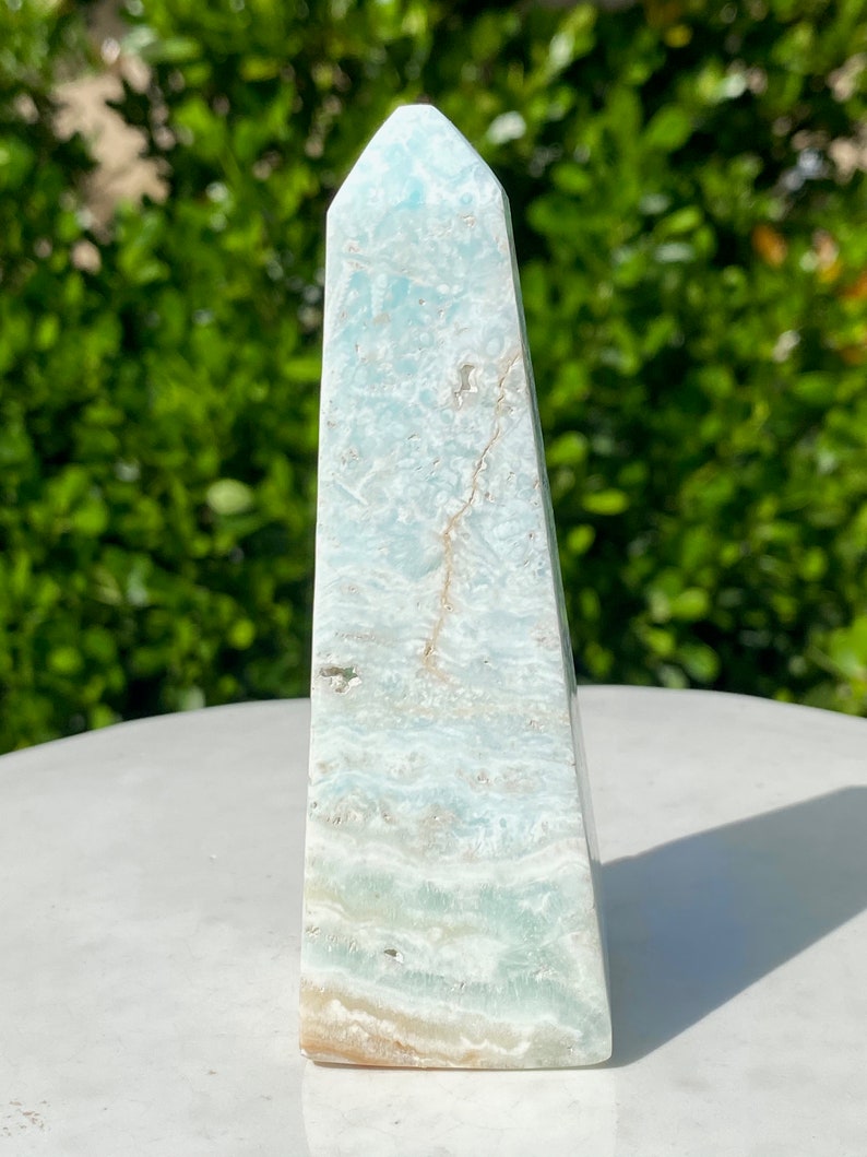 May include: A light blue and white celestite crystal point, also known as a tower or obelisk, with a pointed top and a flat base. The crystal has a natural, raw, and unpolished finish.