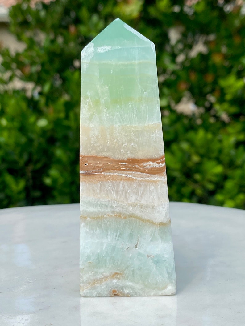 May include: A green, white, and brown banded obelisk crystal point. The crystal is polished and has a smooth, glassy surface.