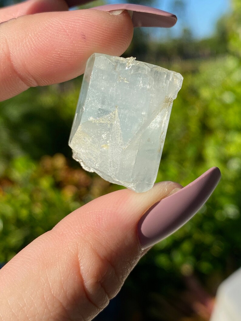 May include: A clear, light blue aquamarine crystal with a rectangular shape. The crystal has a smooth, polished surface and is translucent.