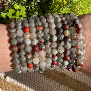 Pu&ograve; includere: Un set di 10 braccialetti realizzati con perline di pietre preziose verdi, rosse e bianche. I braccialetti sono impilati sul polso di una persona.
