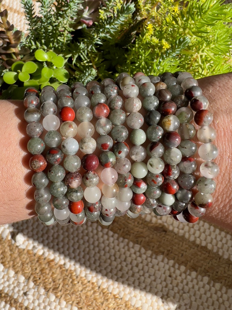 Pu&ograve; includere: Un set di 10 braccialetti realizzati con perline di pietre preziose verdi, rosse e bianche. I braccialetti sono impilati sul polso di una persona.