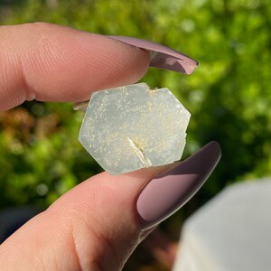 May include: A light blue aquamarine crystal with a hexagonal shape and a white, sparkly inclusion. The crystal is held in a hand.