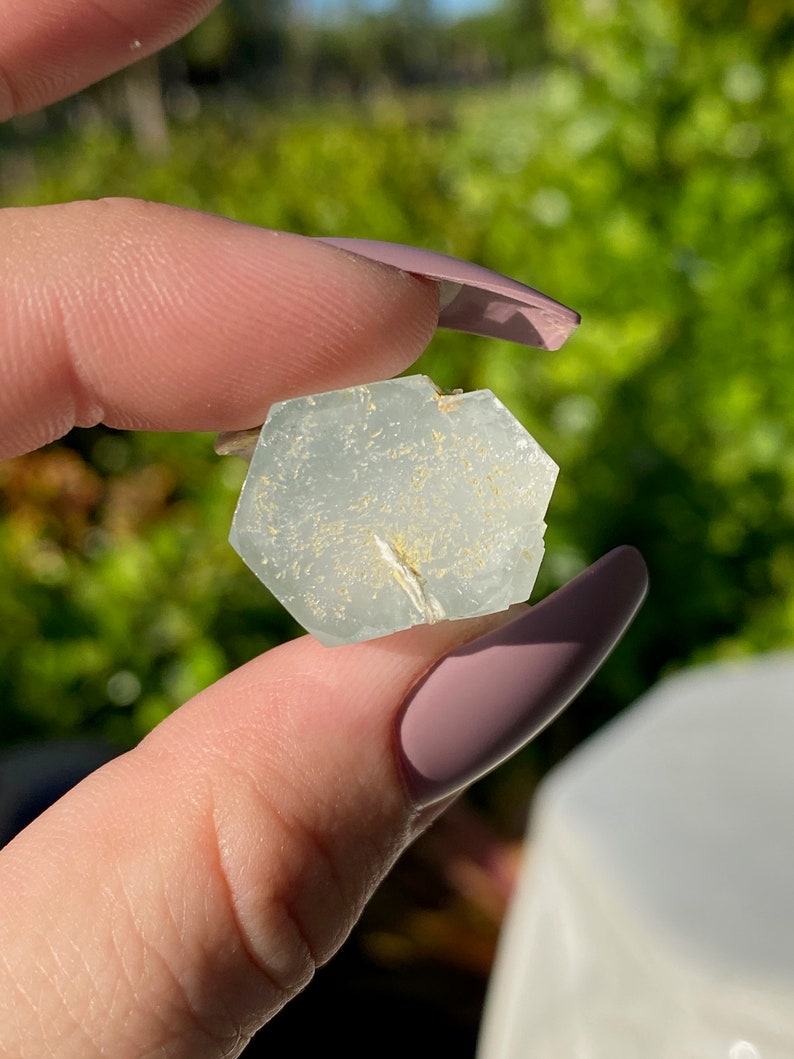 May include: A light blue aquamarine crystal with a hexagonal shape and a white, sparkly inclusion. The crystal is held in a hand.