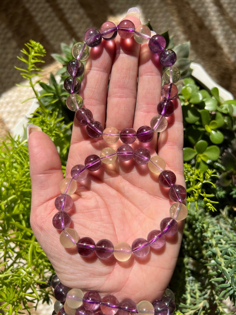 Puede incluir: Dos pulseras hechas con cuentas de piedras preciosas moradas y amarillas. Las pulseras est&aacute;n apiladas una encima de la otra en la mu&ntilde;eca de una persona.
