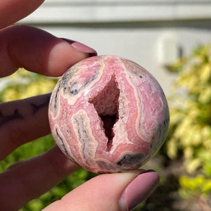 May include: A pink and white banded sphere with a natural crystal cave opening. The sphere is polished and has a smooth surface.