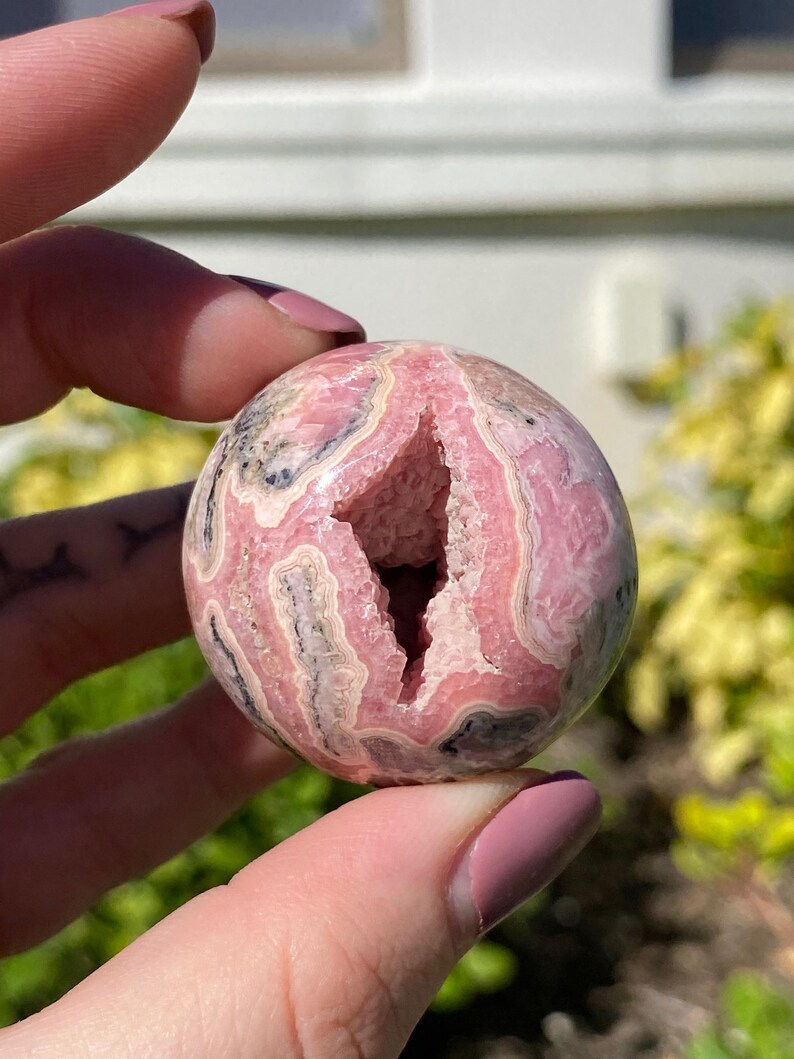 May include: A pink and white banded sphere with a natural crystal cave opening. The sphere is polished and has a smooth surface.
