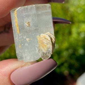 May include: A clear, light blue aquamarine crystal with a natural, raw, and rough texture. The crystal has a rectangular shape and is accented with a small, white, and gold-colored mineral cluster.