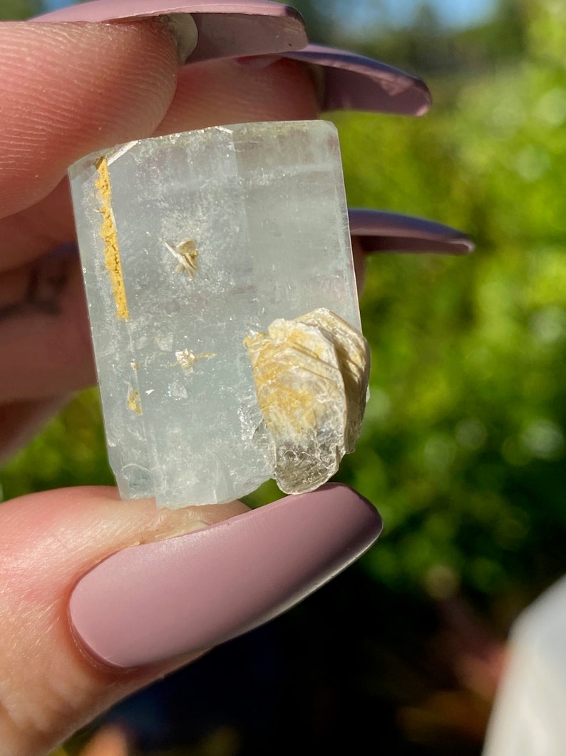 May include: A clear, light blue aquamarine crystal with a natural, raw, and rough texture. The crystal has a rectangular shape and is accented with a small, white, and gold-colored mineral cluster.
