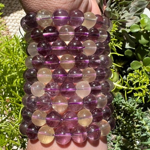 Puede incluir: Un brazalete hecho de cuentas de piedras preciosas moradas y amarillas. Las cuentas son redondas y tienen un acabado liso y pulido.