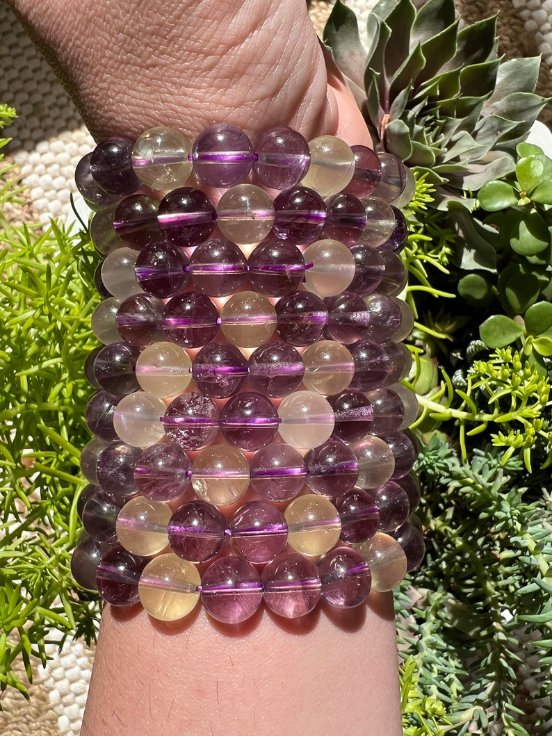 Puede incluir: Un brazalete hecho de cuentas de piedras preciosas moradas y amarillas. Las cuentas son redondas y tienen un acabado liso y pulido.