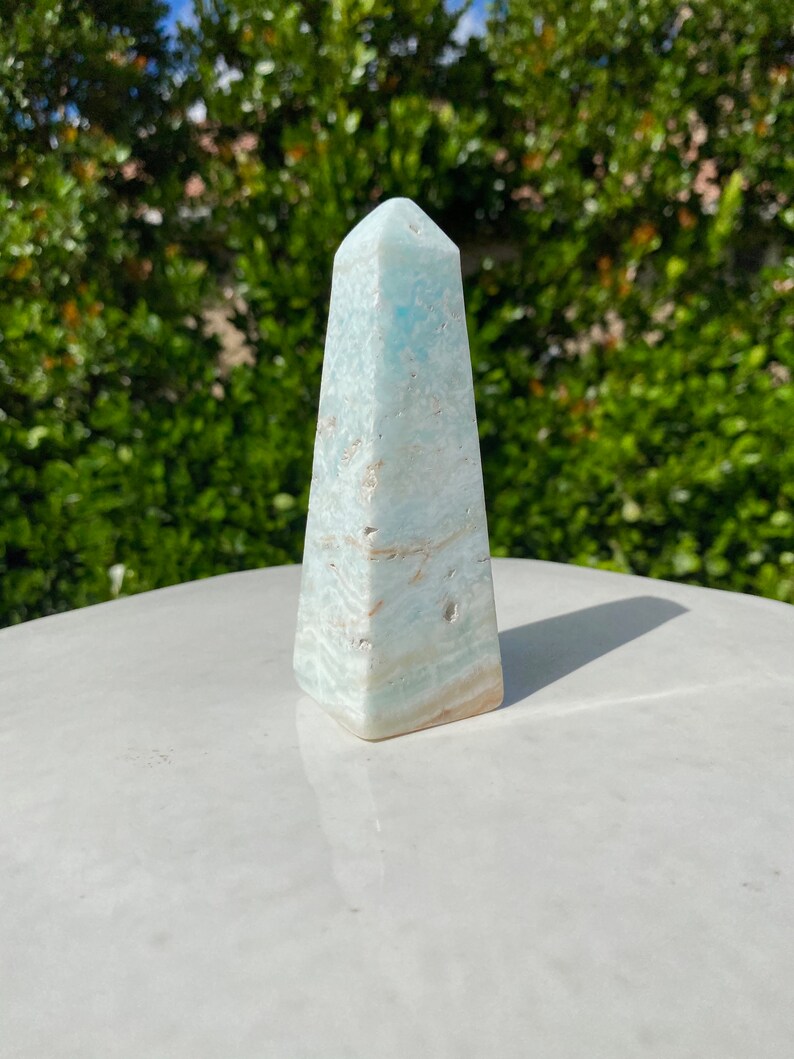 May include: A light blue aquamarine crystal point, also known as an obelisk, with a smooth, polished finish. The crystal is sitting on a white surface.