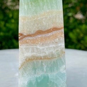 May include: A green, white, and brown layered obelisk crystal. The crystal has a smooth, polished surface.