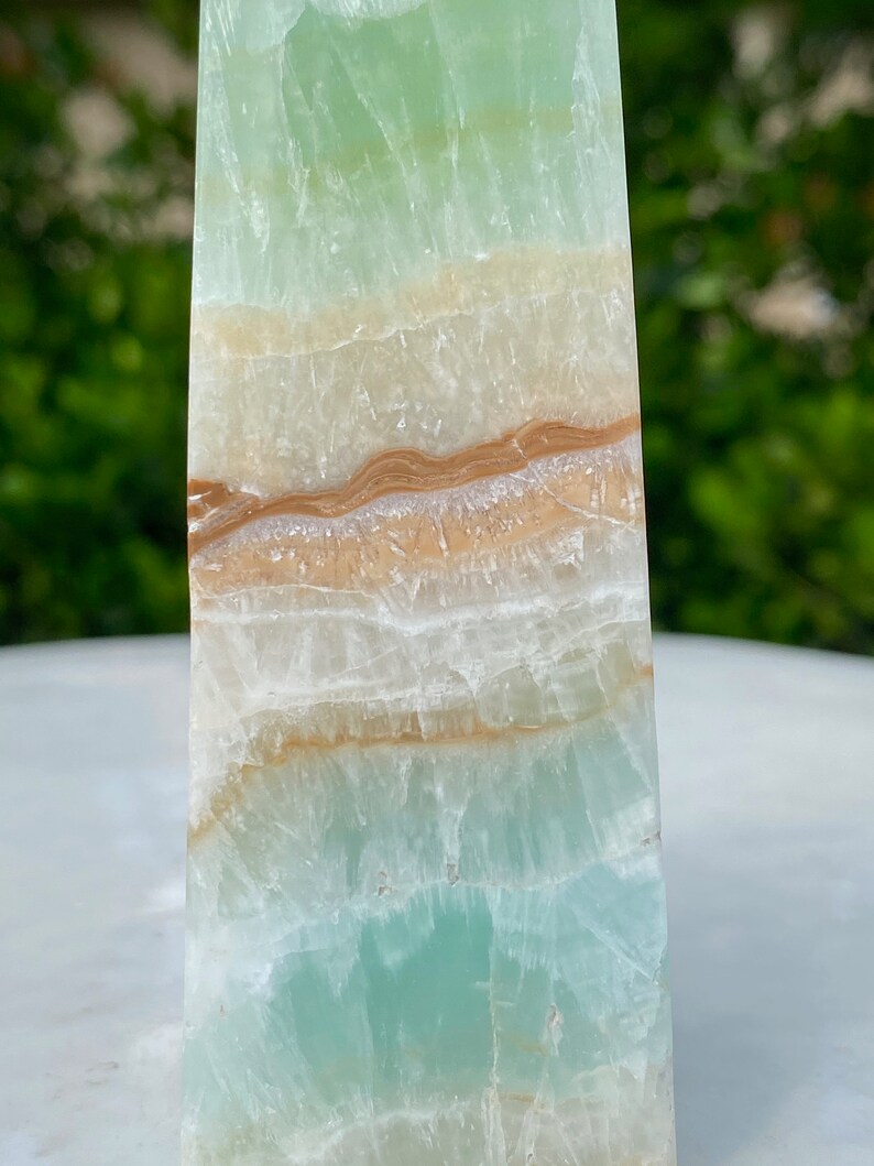 May include: A green, white, and brown layered obelisk crystal. The crystal has a smooth, polished surface.