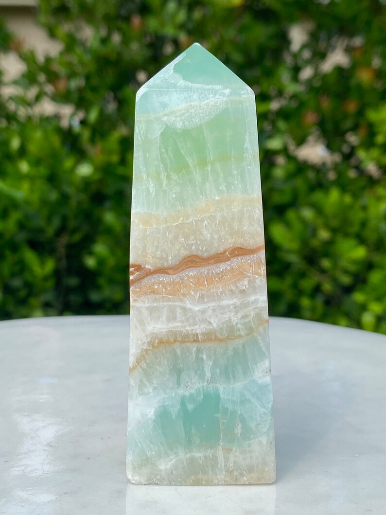 May include: A green and white banded obelisk crystal point with brown and orange banding. The crystal is polished and has a smooth, glassy surface.