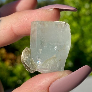 May include: A light blue aquamarine crystal with a clear, glassy texture. The crystal has a rectangular shape with sharp edges and a slightly rounded top. The crystal is held in a hand, and the background is blurred.