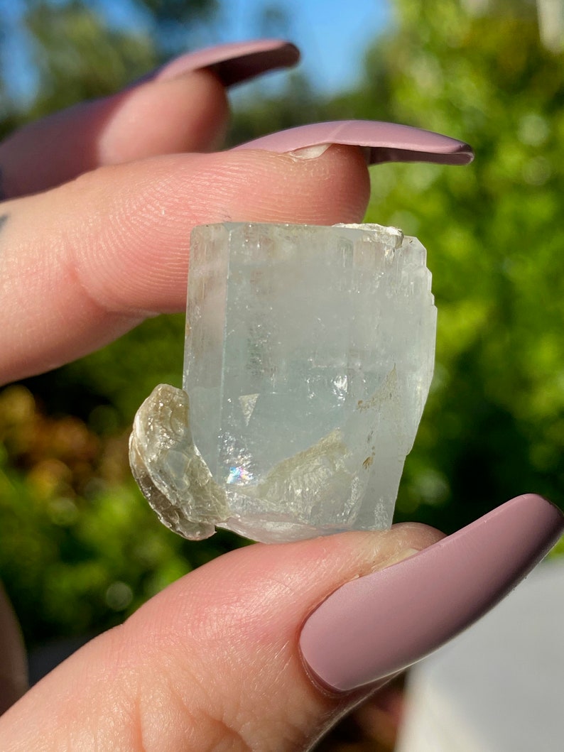 May include: A light blue aquamarine crystal with a clear, glassy texture. The crystal has a rectangular shape with sharp edges and a slightly rounded top. The crystal is held in a hand, and the background is blurred.
