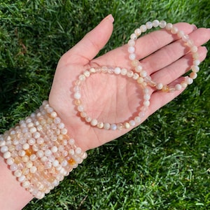 Puede incluir: Dos pulseras hechas con cuentas de piedras preciosas blancas y amarillas. Las pulseras están apiladas en la muñeca de una persona.