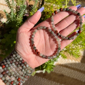 Pu&ograve; includere: Due braccialetti fatti di perline verdi, rosse e trasparenti. I braccialetti sono impilati uno sopra l'altro e sono tenuti da una mano.