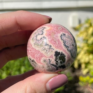 May include: A pink and black sphere of polished rhodochrosite, a type of mineral, with swirling patterns and a smooth surface.