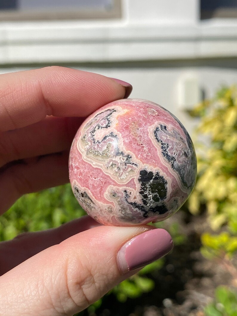 May include: A pink and black sphere of polished rhodochrosite, a type of mineral, with swirling patterns and a smooth surface.