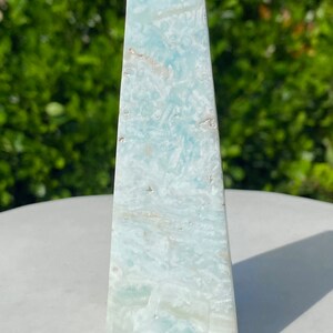 May include: A light blue, white and green celestite obelisk crystal point. The crystal is smooth and polished, with a pointed top and a flat base.