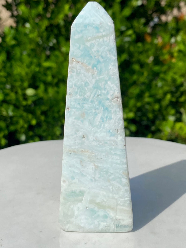 May include: A light blue, white and green celestite obelisk crystal point. The crystal is smooth and polished, with a pointed top and a flat base.