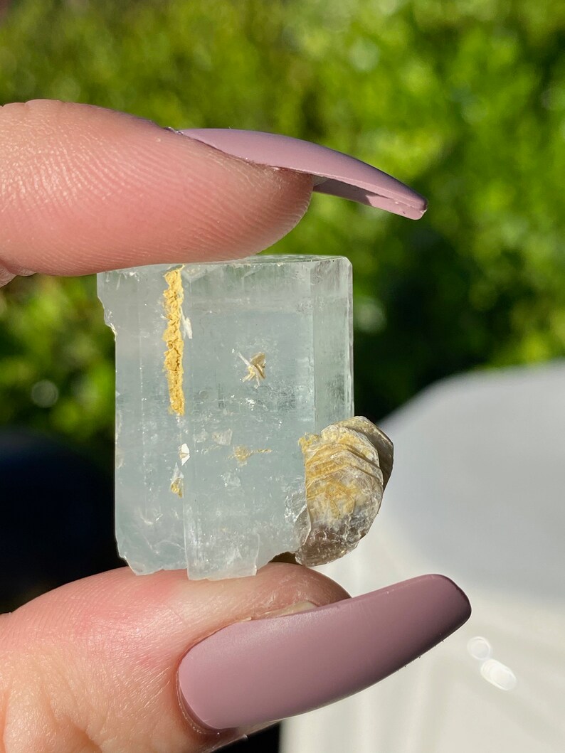 May include: A light blue aquamarine crystal with a natural, raw, and rough texture. The crystal has a clear, glassy appearance and is accented with a small piece of brown mica.