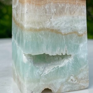 May include: A green, white, and brown layered obelisk crystal with a natural, rough texture. The crystal has a unique, irregular shape and a natural, raw appearance.