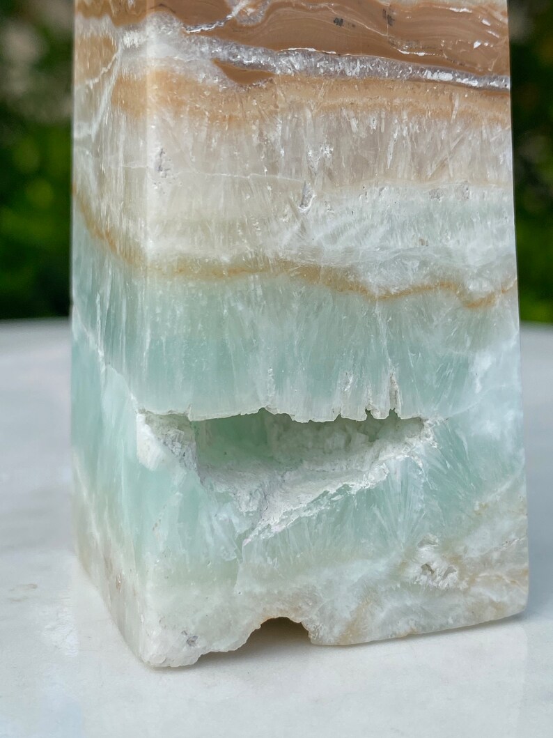 May include: A green, white, and brown layered obelisk crystal with a natural, rough texture. The crystal has a unique, irregular shape and a natural, raw appearance.