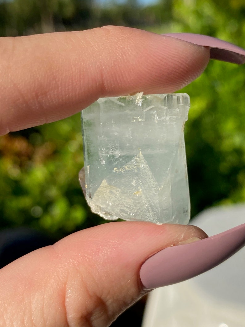 May include: A clear, light blue aquamarine crystal with a slightly cloudy appearance. The crystal is faceted and has a rectangular shape.