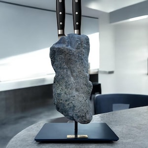 Könnte beinhalten: Ein einzigartiger Messerblock mit zwei Messern mit schwarzen Griffen, die in einen strukturierten, grauen Stein gesteckt sind. Der Stein ist auf einem schwarzen Sockel mit einer kleinen goldfarbenen Plakette montiert. Die Messer haben silberne Zwingen und drei Nieten an den Griffen.