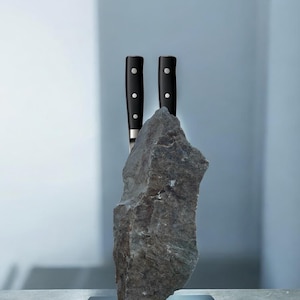 Könnte beinhalten: Ein einzigartiger Messerblock mit einem großen, grauen Stein und zwei eingesetzten Messern. Die Messer haben schwarze Griffe mit drei silbernen Nieten. Der Stein steht auf einem schwarzen rechteckigen Sockel, ein modernes Küchenaccessoire.
