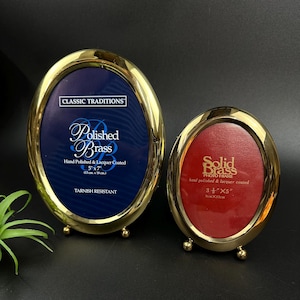 May include: Two oval brass photo frames. The larger frame has a blue interior and reads "Polished Brass" and is 5x7 inches (13cm x 18cm). The smaller frame has a red interior and reads "Solid Brass" and is 3.5 x 5 inches (9cm x 13cm).