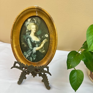 May include: An oval-shaped, antique gold-colored picture frame with a portrait of a woman in a period dress. The frame is supported by a decorative, bronze-colored easel. The portrait features a woman holding a flower.