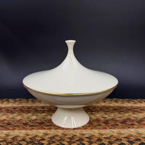 May include: A white and beige lidded serving dish with a gold trim. The dish sits on a pedestal base. The lid has a rounded shape with a small, elongated finial. The dish is likely made of ceramic or porcelain.