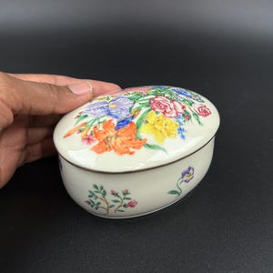 May include: Oval-shaped ceramic trinket box with a floral design. The lid features a vibrant arrangement of colorful flowers, including orange lilies, yellow daffodils, and pink roses. The box has a cream-colored base with a brown trim.