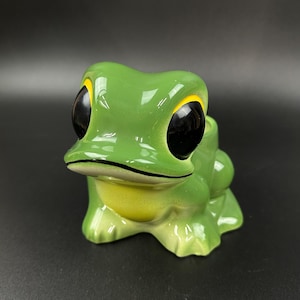 May include: A ceramic frog figurine with large black eyes, yellow accents, and a glossy green finish. The frog has a light green belly and is sitting on a dark surface. This decorative item is a whimsical addition to any home decor.