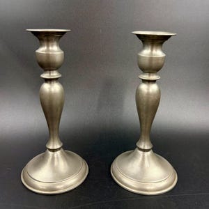 May include: Two antique silver-toned candlesticks. Each candlestick has a wide, circular base, a slender, elongated stem, and a cup-shaped top. The candlesticks are made of metal and have a classic, elegant design. They are displayed against a dark background.