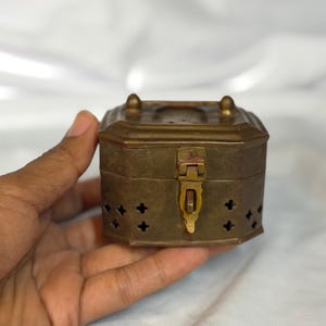 May include: A small, octagonal brass box with a hinged lid and a central metal latch. The sides of the box feature a repeating pattern of small, cross-shaped cutouts. The top surface has a raised border and two small, rounded decorative knobs.