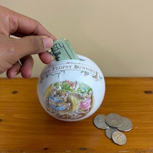 May include: A white ceramic sphere-shaped piggy bank with a colorful illustration of rabbits and the text "The Flopsy Bunnies." The bank has a slot at the top and is filled with paper money. Several coins are on the wooden surface.