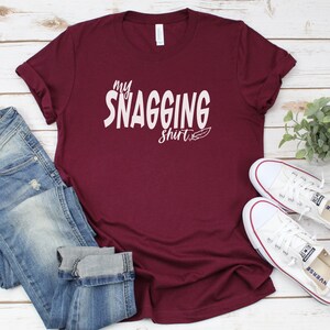 May include: A maroon t-shirt with the text "my SNAGGING shirt" in white lettering. The shirt is laid flat on a white wooden surface with a pair of white sneakers and blue jeans.